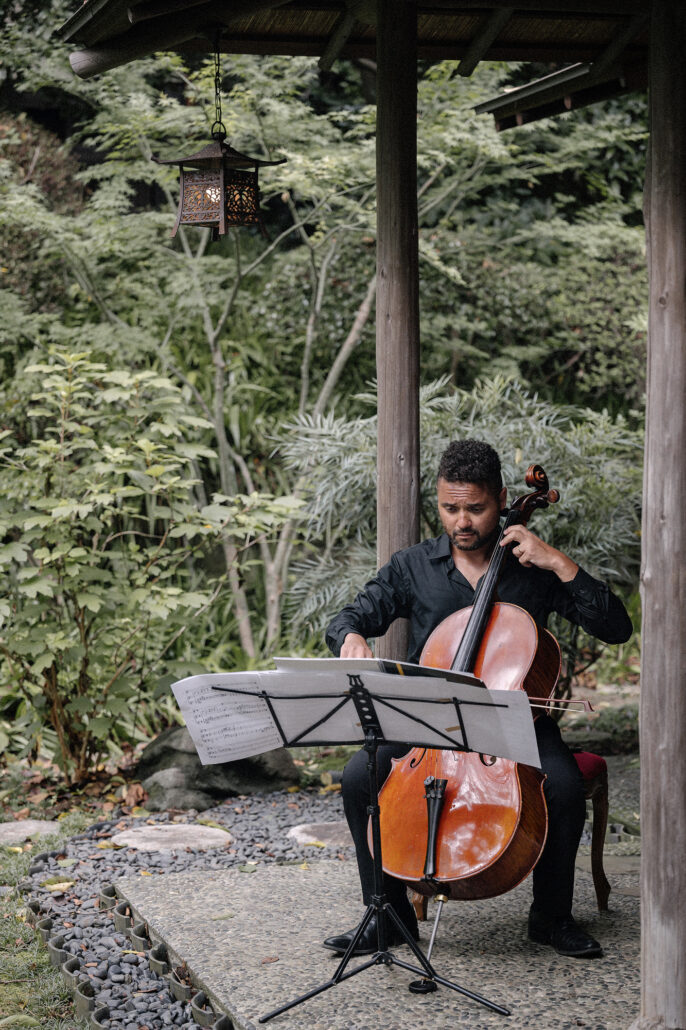A cellist plays under a wooden garden pavilion with a hanging lantern, music for a Japanese garden elopement ceremony.