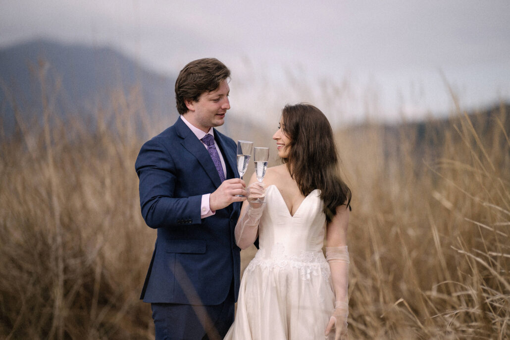 The groom and bride clink champagne glasses among tall golden grass with misty hills behind, a champagne toast at a Japan elopement.