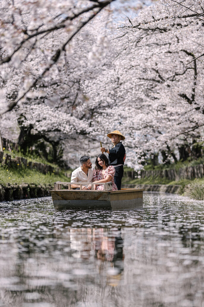 A boatman poles the couple along a narrow river beneath arching cherry blossom.