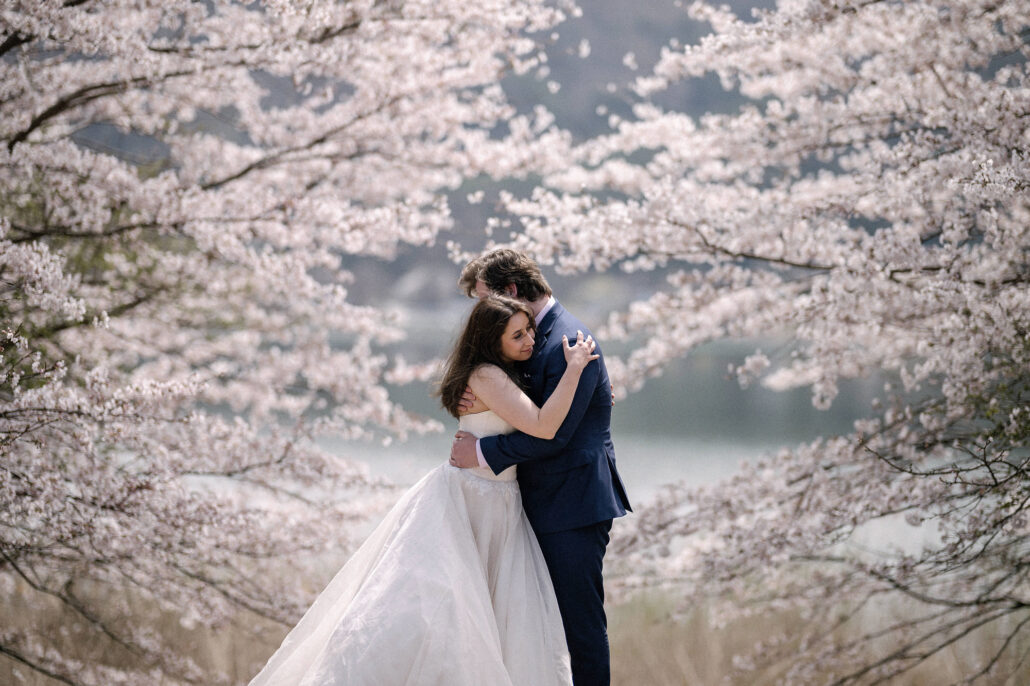 The bride and groom hold each other inside a frame of cherry blossom with a lake behind them, a cherry blossom elopement in Japan.