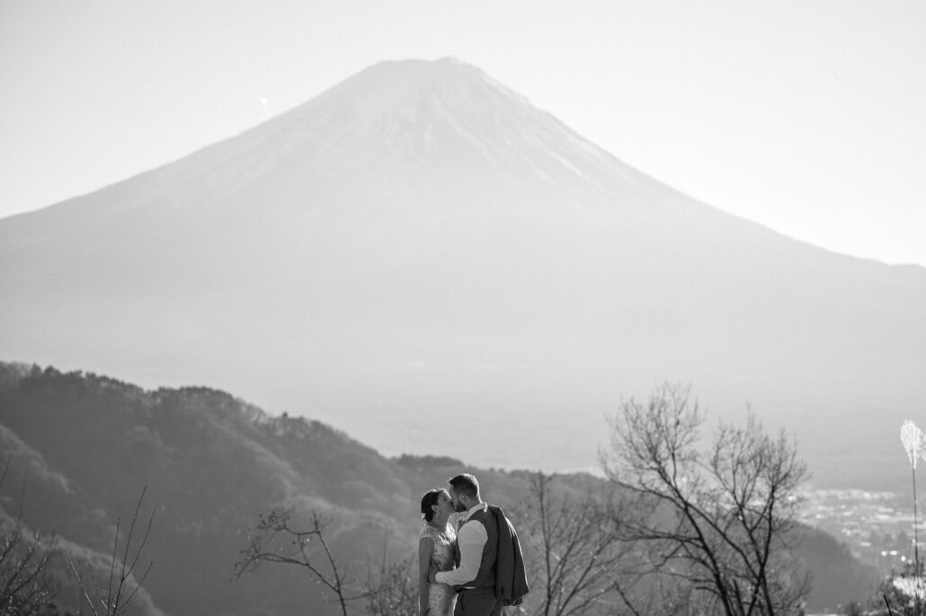 The couple embrace on the hillside below Mt Fuji in hazy winter light, bare branches around them, near the end of their elopement.