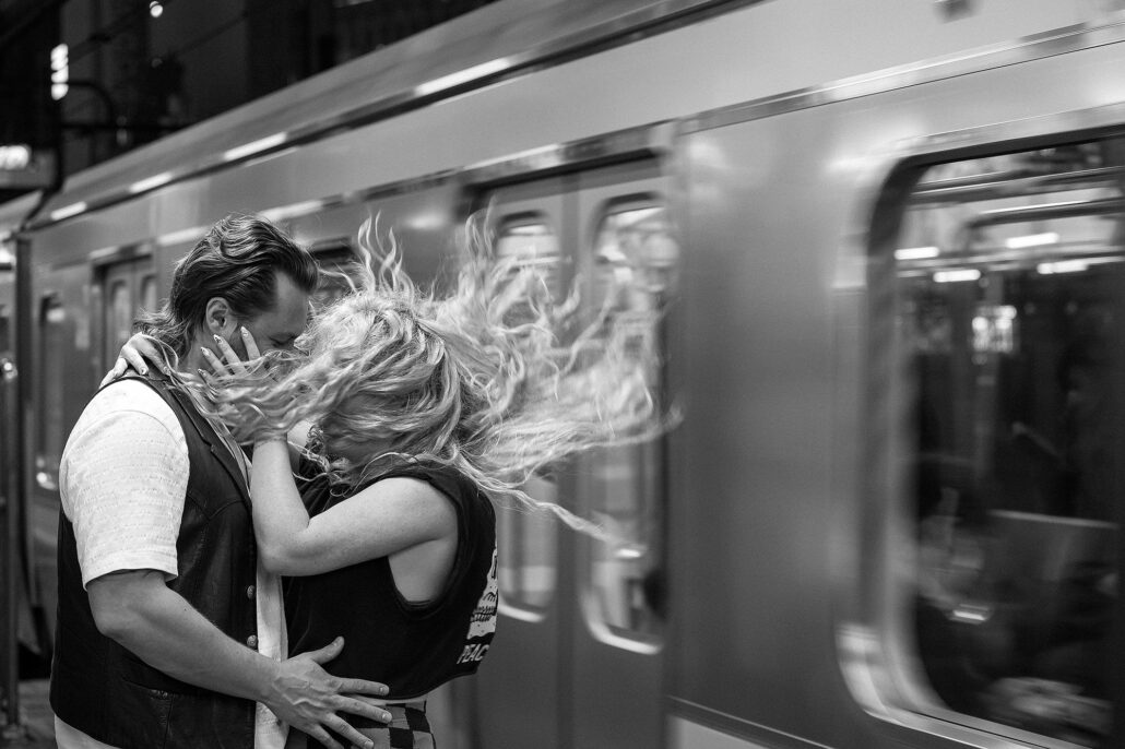 In black and white, a couple kiss on a station platform as a train rushes past, her hair flying in its wind.