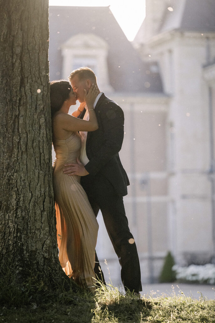 The bride leans against a tree trunk as the groom kisses her in the golden light outside a chateau in France.