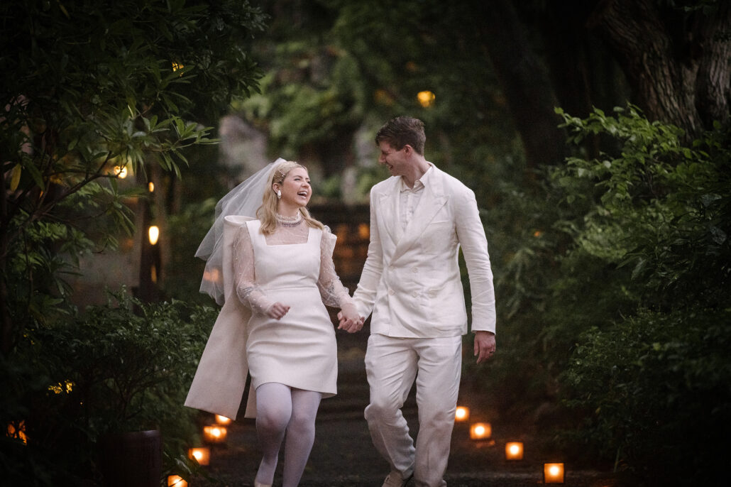 The bride and groom laugh as they walk hand in hand down a lantern-lit path through the ryokan garden at Tsurumaki Onsen.