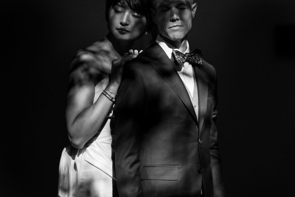 In black and white, the bride rests her hands on the groom's shoulders as light falls across them in stripes.