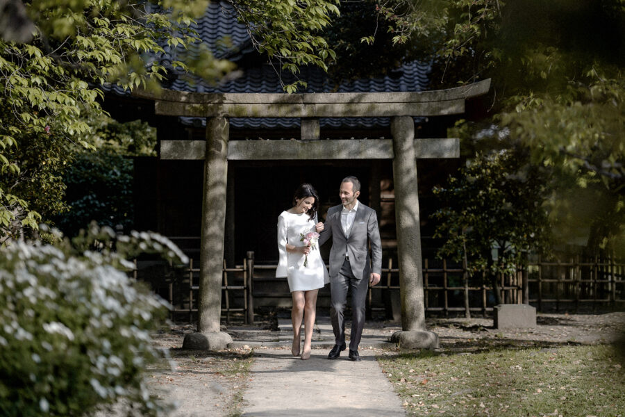 A couple walk arm in arm beneath a weathered stone torii in a Japanese garden in spring, a small pink bouquet in her hand.