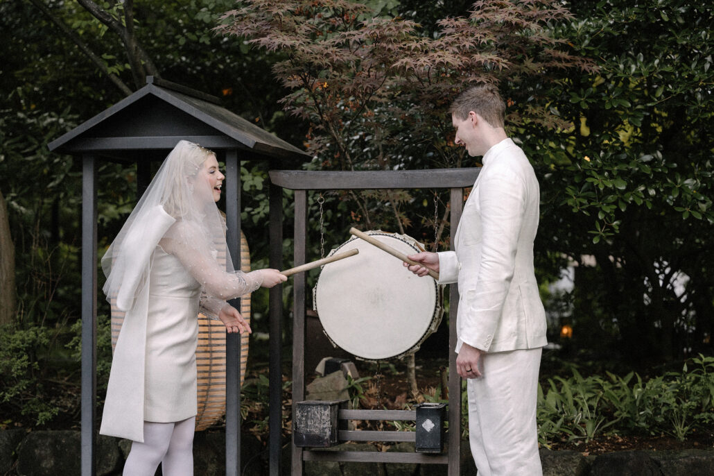 The bride and groom strike a taiko drum together in the ryokan garden, laughing beneath red maple leaves after their elopement.