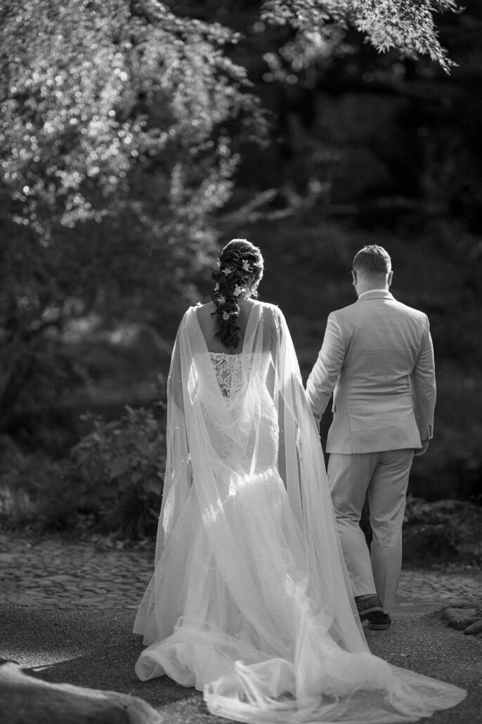 The bride and groom walk away along a garden path in Tokyo, her lace cape trailing, in black and white.