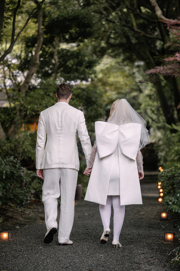 The couple walk hand in hand away down a lantern-lit path through the trees, her veil and dress bow white against the dusk.