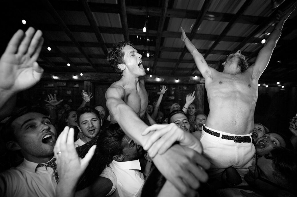 In black and white, two shirtless men are lifted high above a cheering crowd on the dance floor at a wedding in Montana.
