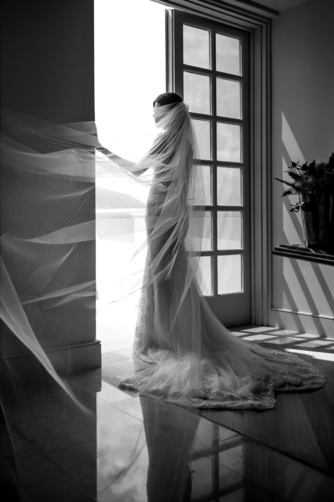 In black and white, the bride stands at a tall window as her long veil drifts across the room, getting ready for her destination wedding in Cyprus.