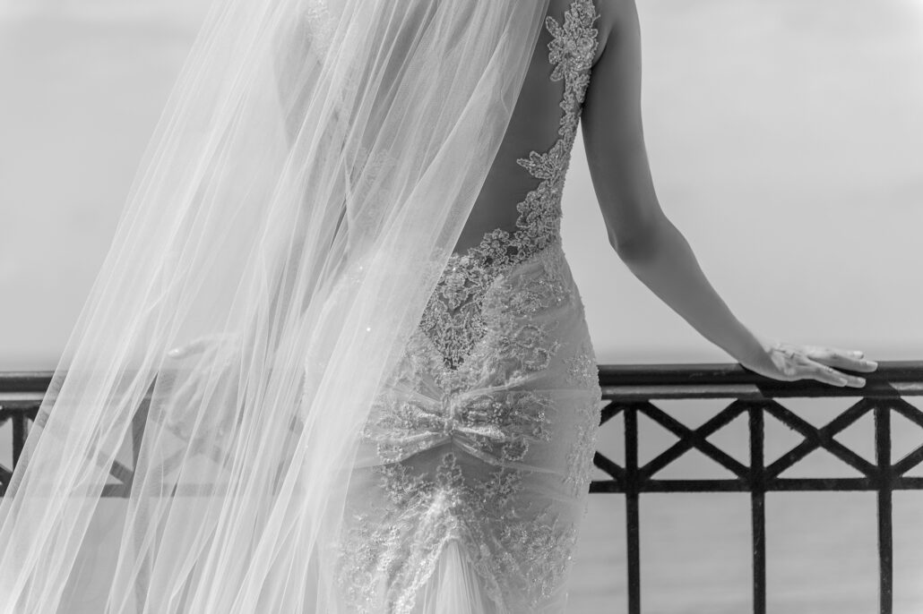 In black and white, the bride rests her hand on a balcony rail above the sea, her veil drifting over the lace of her gown.