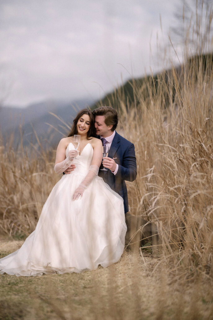 The bride and groom sit close in tall grass holding champagne glasses after their vows, an elopement toast near Mt Fuji.