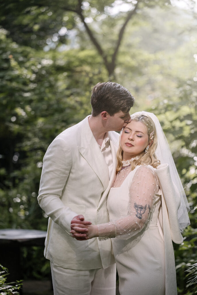 The groom kisses the bride's forehead as she closes her eyes, an embrace among the trees during their elopement in Japan.