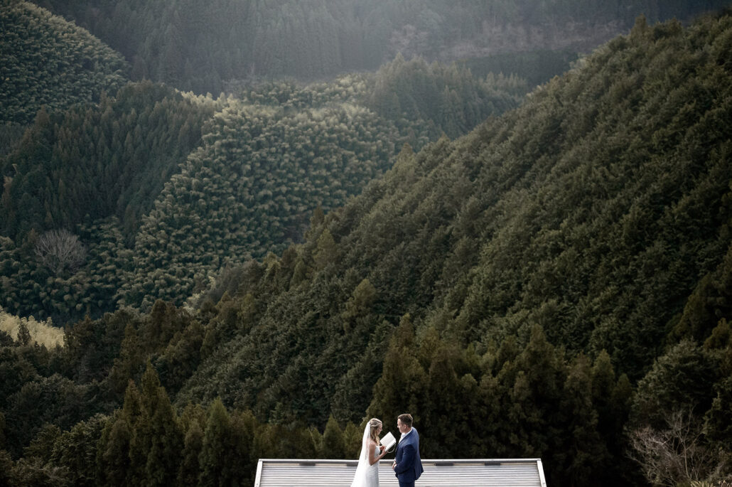 A couple stand small on a terrace below steep forested mountains, deep in the countryside of Japan.