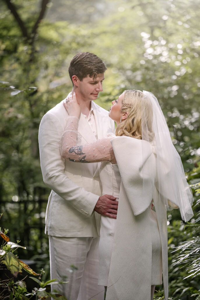 The bride's hand rests on the groom's face beneath tall trees, elopement photography in a forest garden near Tokyo.