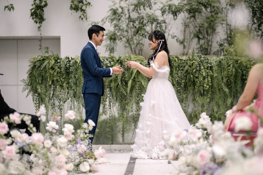 The couple exchange rings beside a wall of hanging ferns, pink and white flowers in front, at an Eneko Tokyo wedding in Nishi-Azabu.