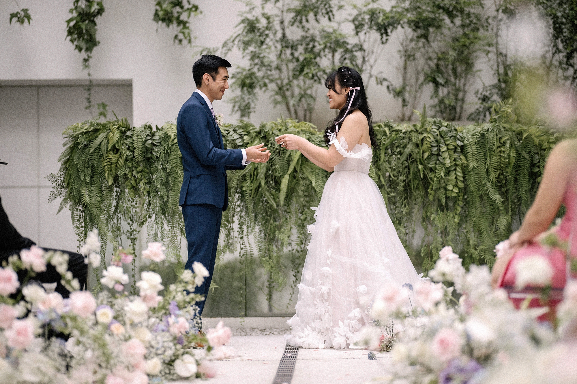 The couple exchange rings beside a wall of hanging ferns, pink and white flowers in front, at an Eneko Tokyo wedding in Nishi-Azabu.