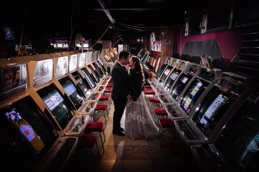 A couple in wedding clothes kiss between rows of retro arcade cabinets, wedding photos at Game Center Mikado in Takadanobaba.