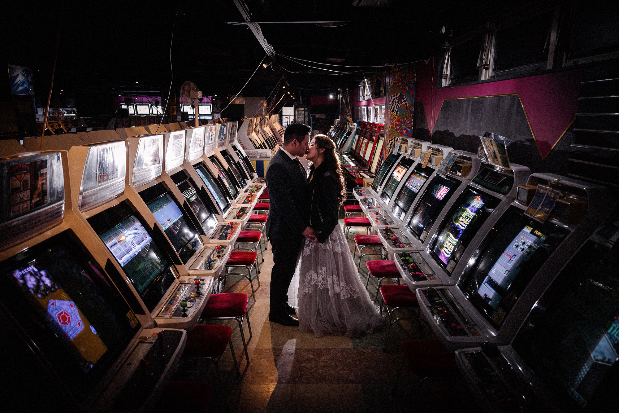 A couple in wedding clothes kiss between rows of retro arcade cabinets, wedding photos at Game Center Mikado in Takadanobaba.