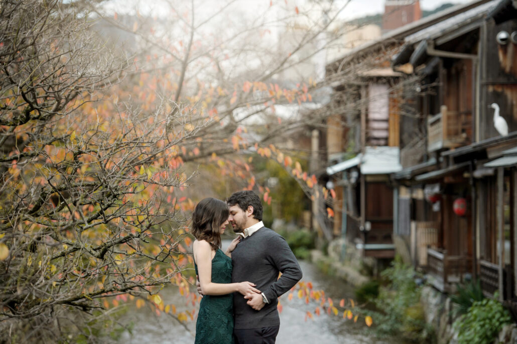 Away from the crowds in Gion: a couple embrace beside the Shirakawa canal in autumn, old wooden teahouses behind them.