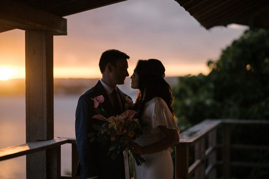 The bride and groom kiss on the deck at Hyakuna Garan as the sun sets behind them, her bouquet of soft roses held between them.