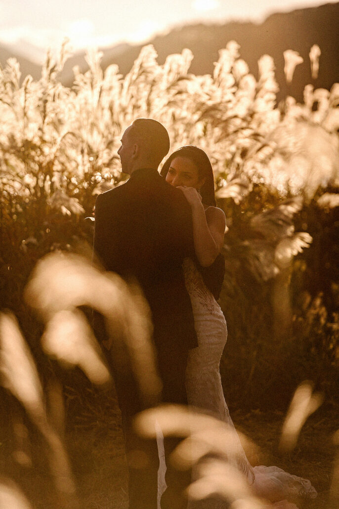 The bride looks out from behind the groom's shoulder, framed by glowing pampas grass at golden hour during wedding photos near Mt Fuji.