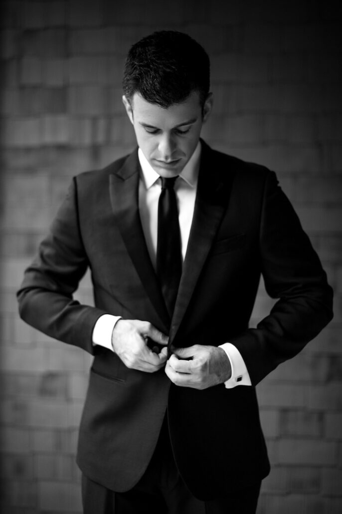 In black and white, a groom fastens the button of his suit jacket on the morning of his wedding.