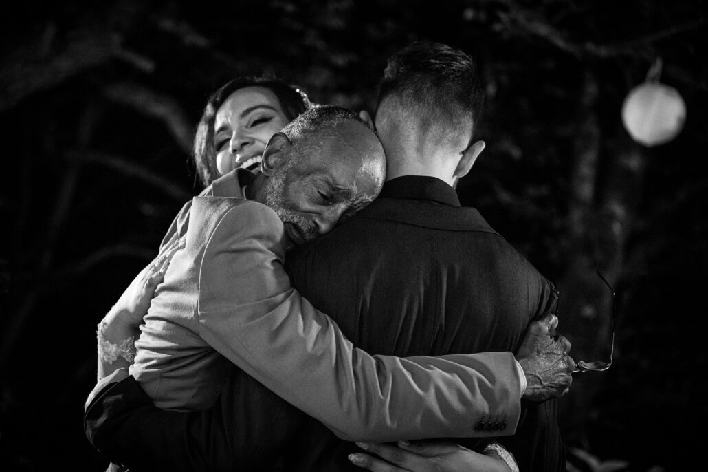 In black and white, an older guest embraces the groom while the bride laughs over their shoulders.