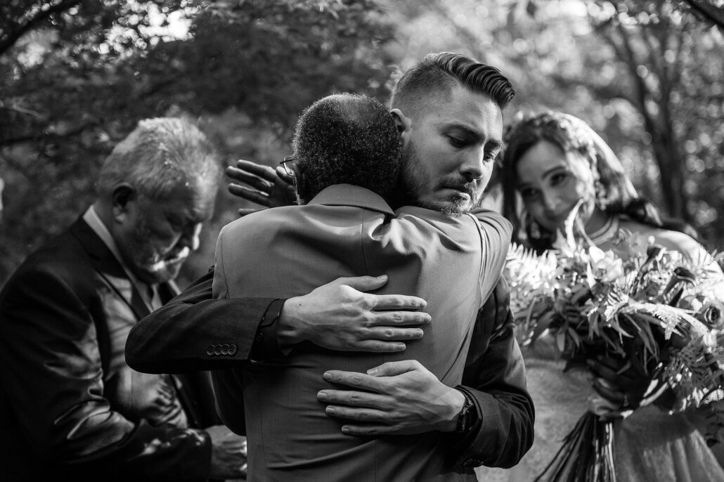 In black and white, the groom holds an older man in a long embrace, eyes closed, as the bride looks on with her bouquet.