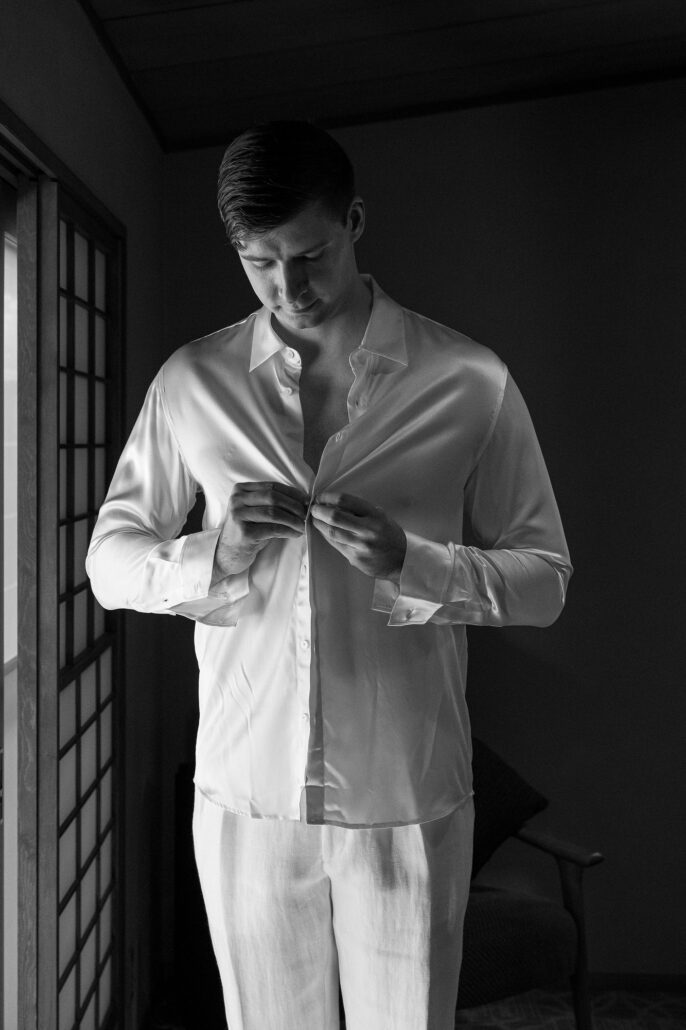 A black and white portrait of the groom getting ready by a shoji screen, fastening his shirt before the elopement ceremony.
