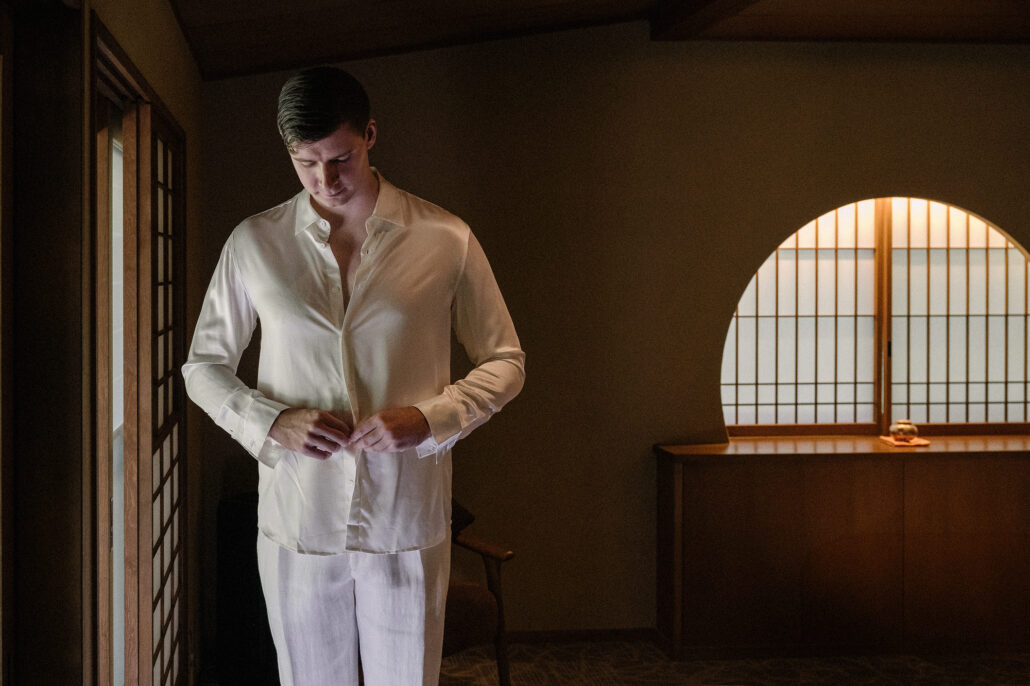The groom buttons his ivory shirt beside a paper window in a traditional Japanese inn, getting ready for his elopement.