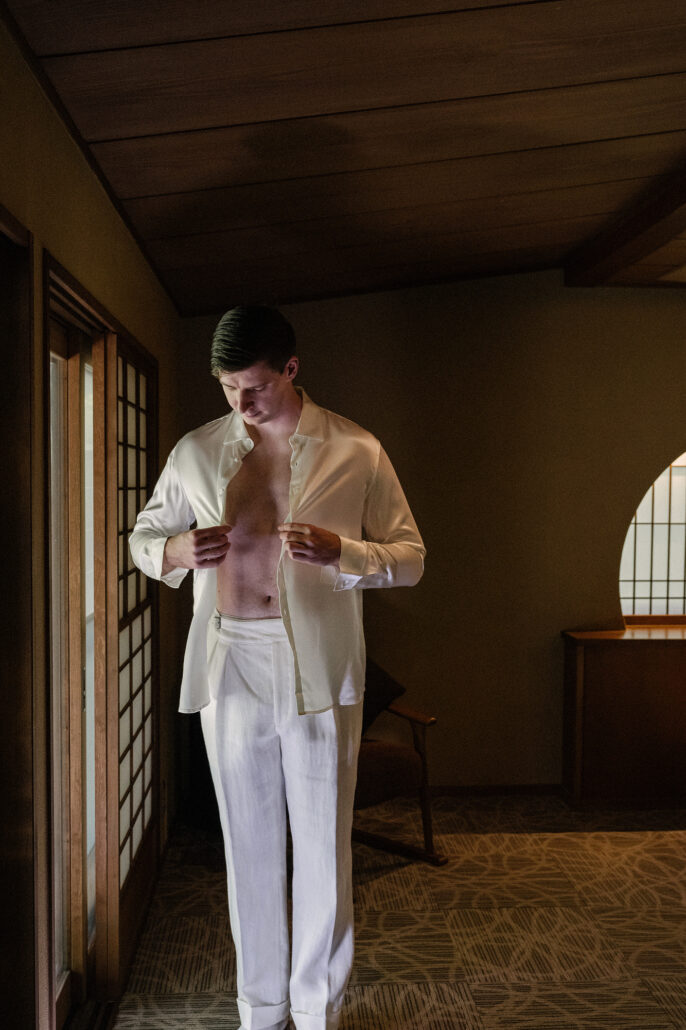 The groom stands in a traditional ryokan room with his shirt open, getting ready for the elopement in ivory trousers and shirt.