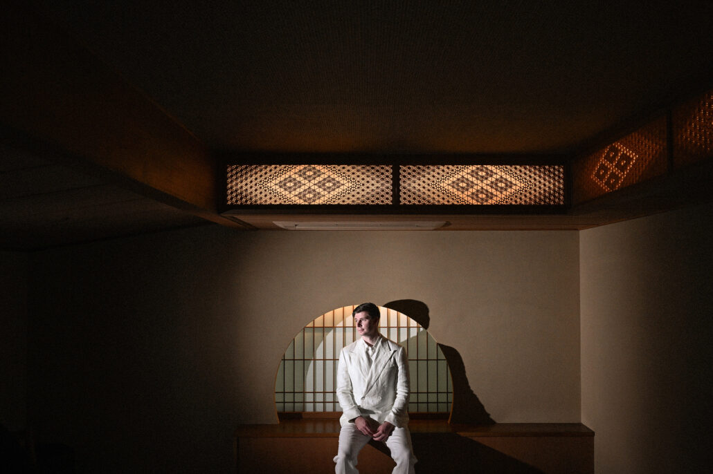 The groom sits in a round shoji window in a dark wooden ryokan room, getting ready for an elopement at Tsurumaki Onsen.