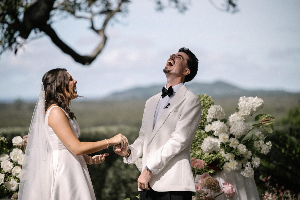 The groom throws his head back laughing as he holds the bride's hands during their vows, white flowers and hills behind them.