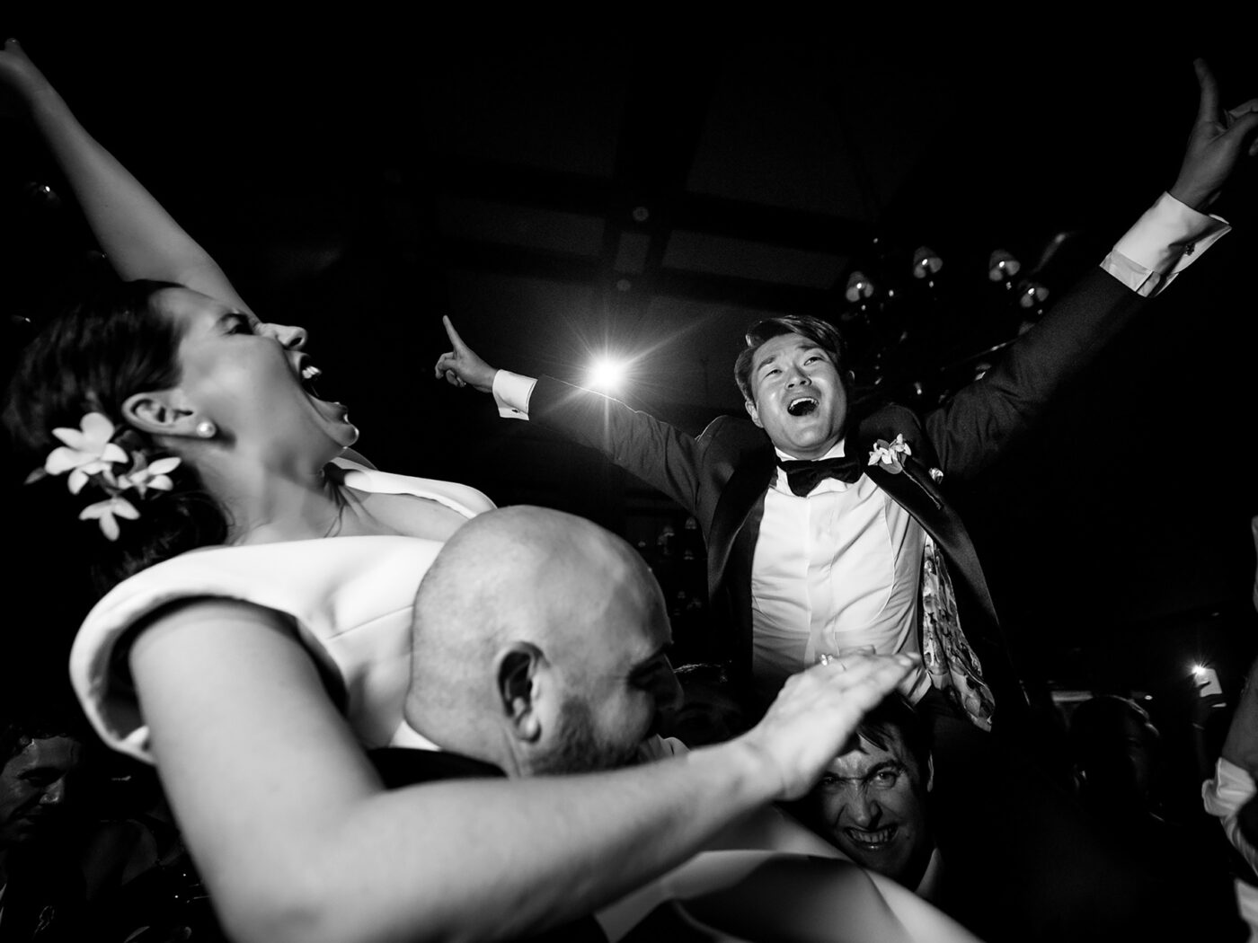 In black and white, the groom is lifted high by his friends, arms flung wide, as the bride shrieks with laughter beside him.