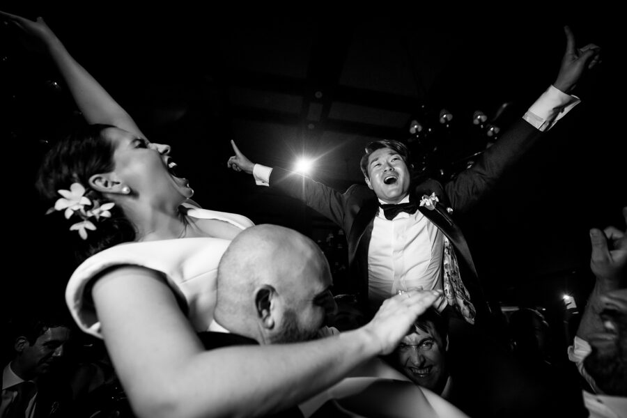 In black and white, the groom is lifted high by his friends, arms flung wide, as the bride shrieks with laughter beside him.