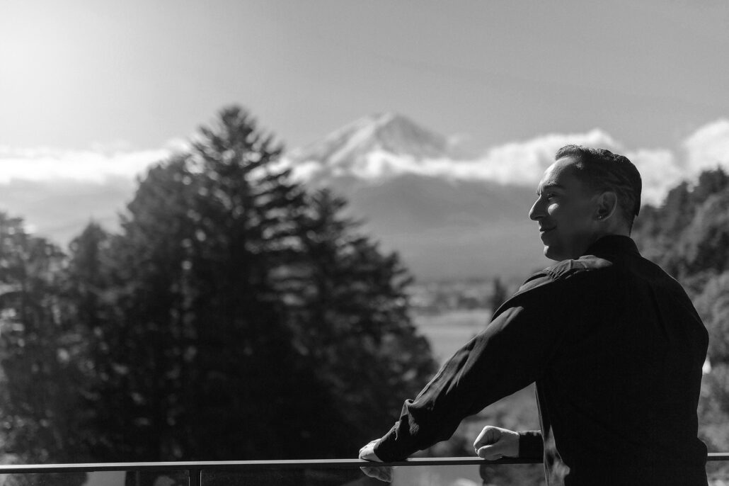 In black and white, the groom leans on a railing and looks out toward Mt Fuji rising above a band of cloud.