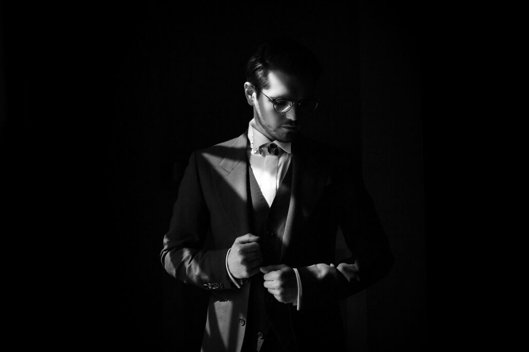 The groom fastens his jacket in a single beam of light, the rest of the room in shadow.