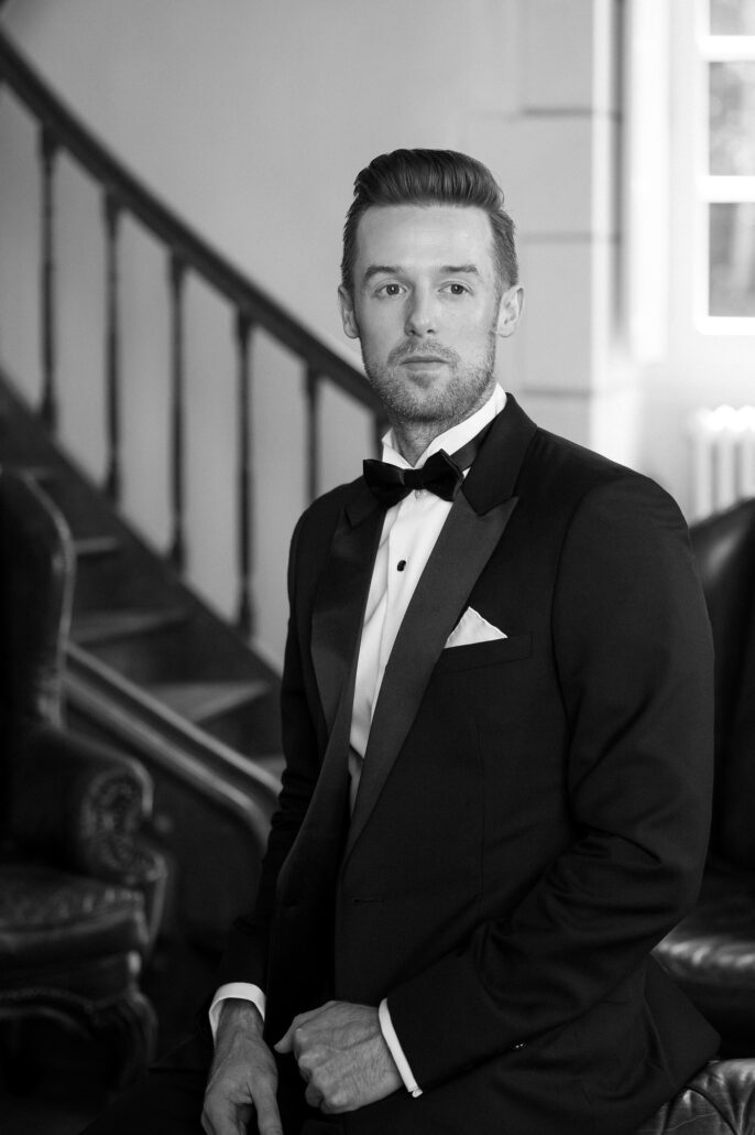 In black and white, the groom sits in his tuxedo by the staircase of an old French house.