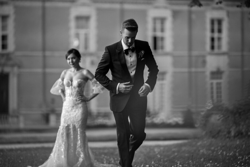 In black and white, the groom strides toward the camera in his tuxedo as the bride follows, a chateau behind them.