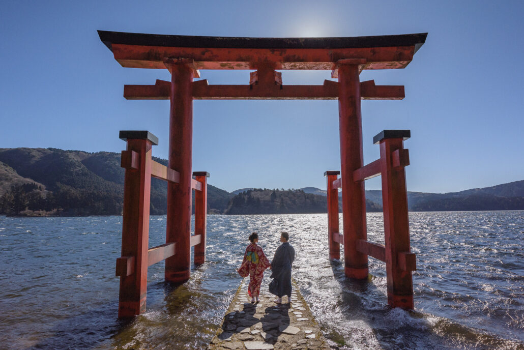 A couple in kimono walk out beneath the vermilion torii that stands in the water of Lake Ashi at Hakone, the lake glittering in the sun.