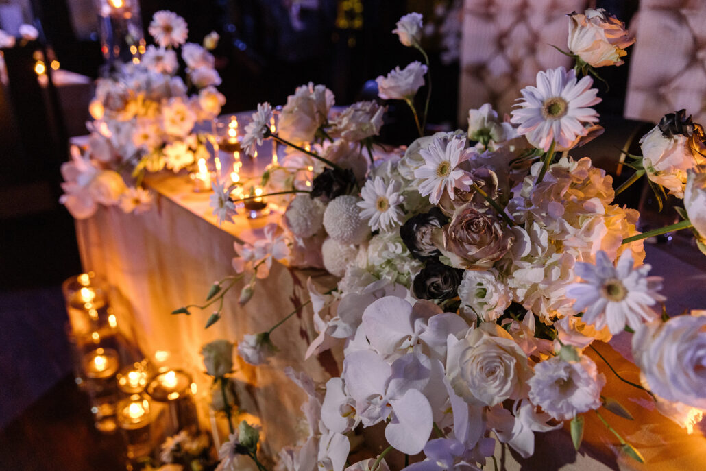 Candlelight warms a bank of white daisies, roses and orchids along the head table at the reception.