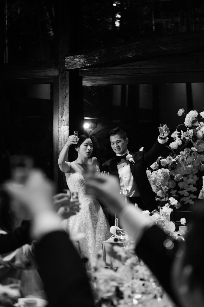 In black and white, the bride and groom raise their glasses from the head table as guests toast them back.