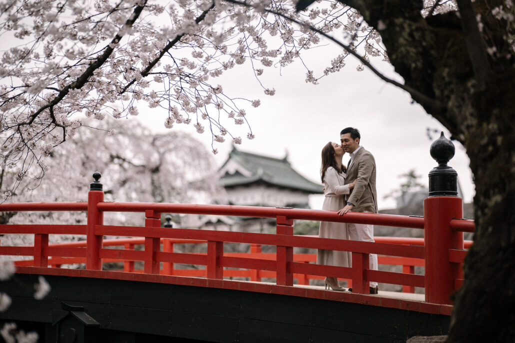 The couple kiss on a red bridge in Hirosaki Park, the castle and cherry blossom behind them.