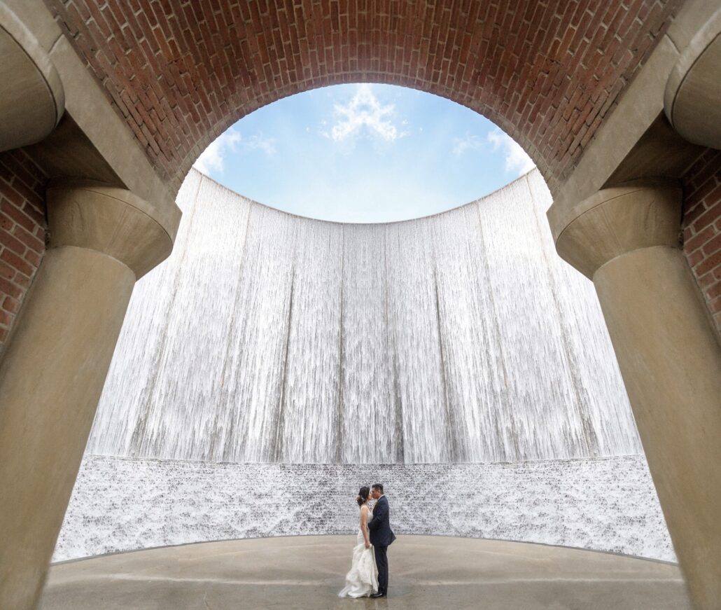 The bride and groom stand small beneath the arch of the Waterwall in Houston, the water falling all around them.