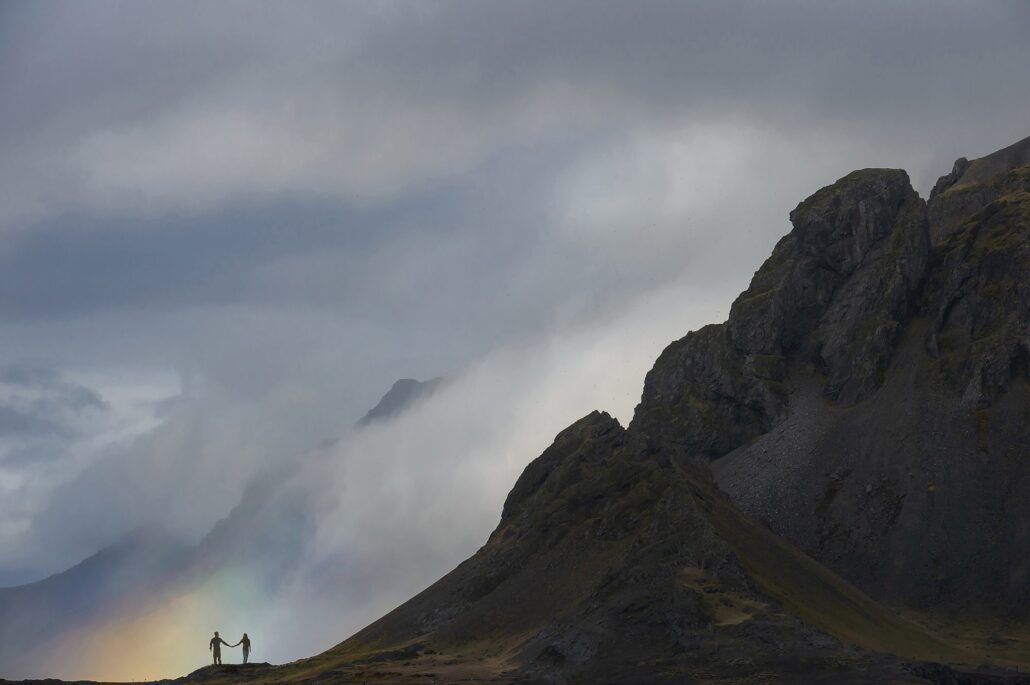 A couple stand tiny on a dark mountainside in Iceland as a rainbow breaks through the cloud below them.