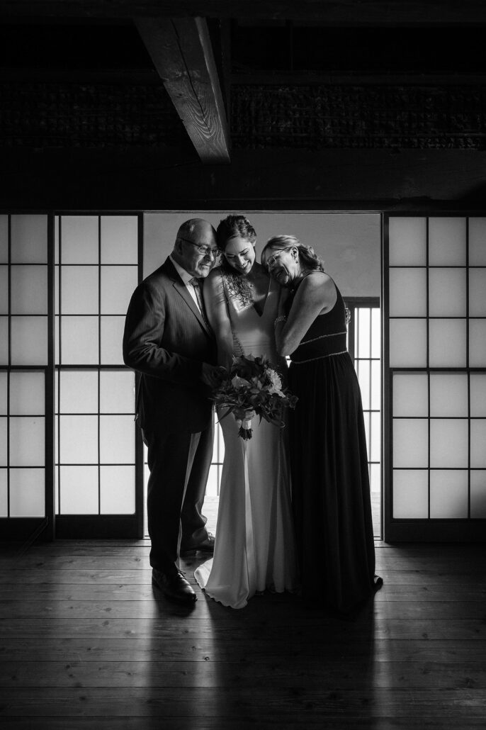 In black and white, an older couple wrap the bride in a hug in a room lit by tall shoji windows.