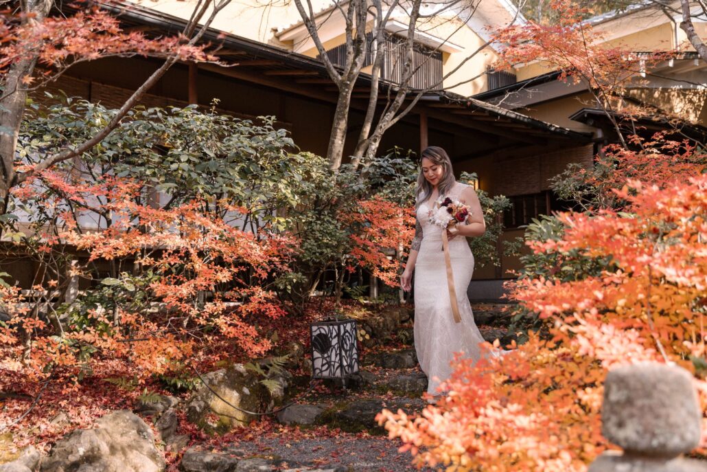 The bride walks into a Kyoto maple garden in late November, red leaves all around her and a garden roof behind.