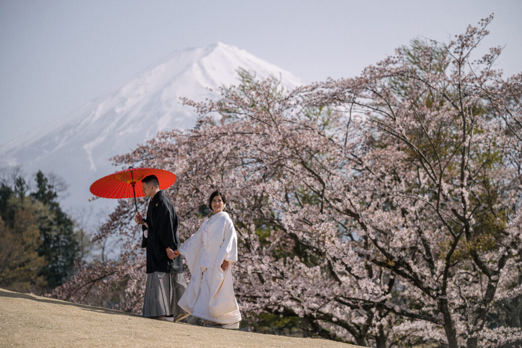 A couple in wedding kimono walk beneath cherry blossom, the groom holding a red umbrella, with snow-capped Mt Fuji behind them at a wedding in Japan.